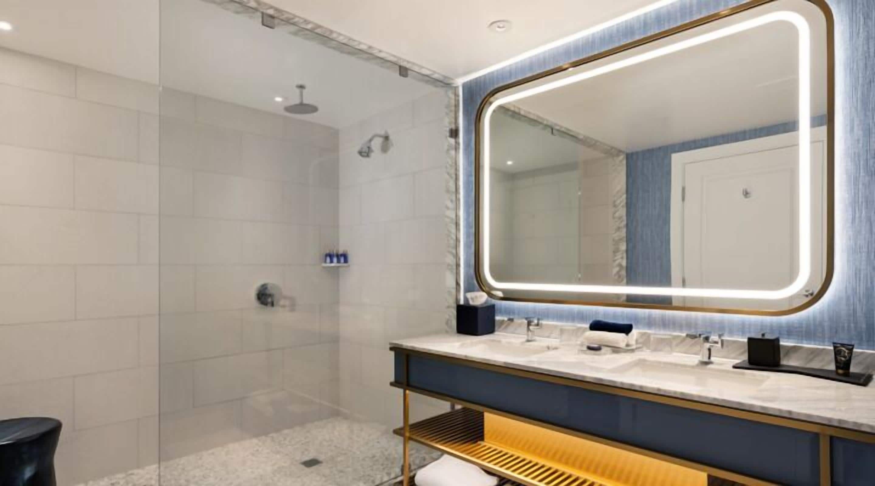Un baño moderno con ducha envuelta en vidrio, un espejo grande con bordes iluminados y un tocador con lavamanos dobles y un perchero para toallas.