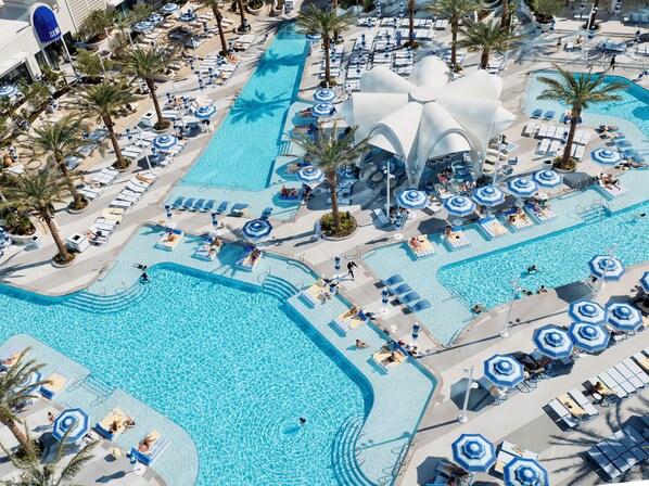 A large outdoor swimming pool with multiple lanes, surrounded by sun loungers and umbrellas, and palm trees.
