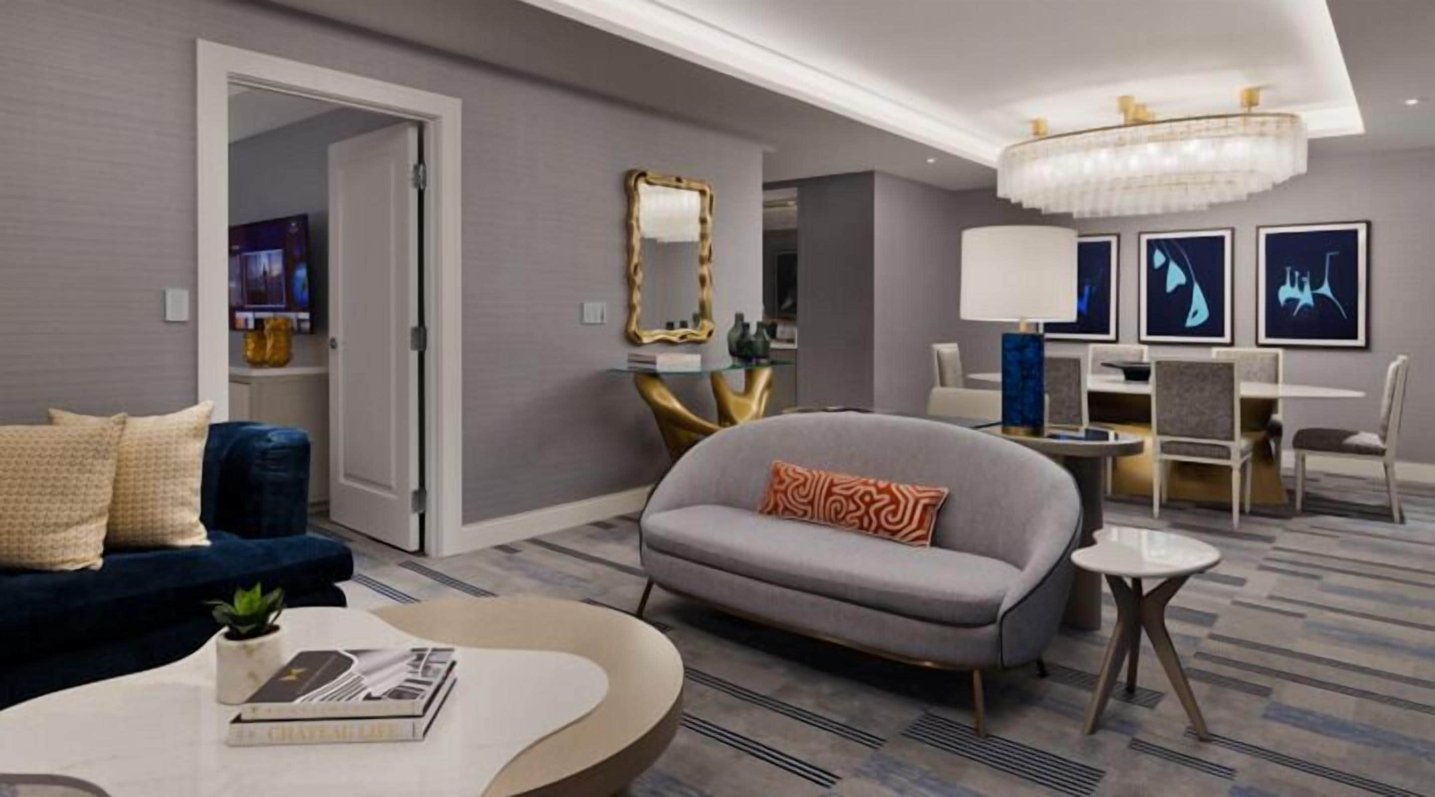 Quarto de hotel moderno com um sofá cinzento, uma mesa de centro redonda, uma área de jantar com uma mesa e cadeiras, e um candelabro.
