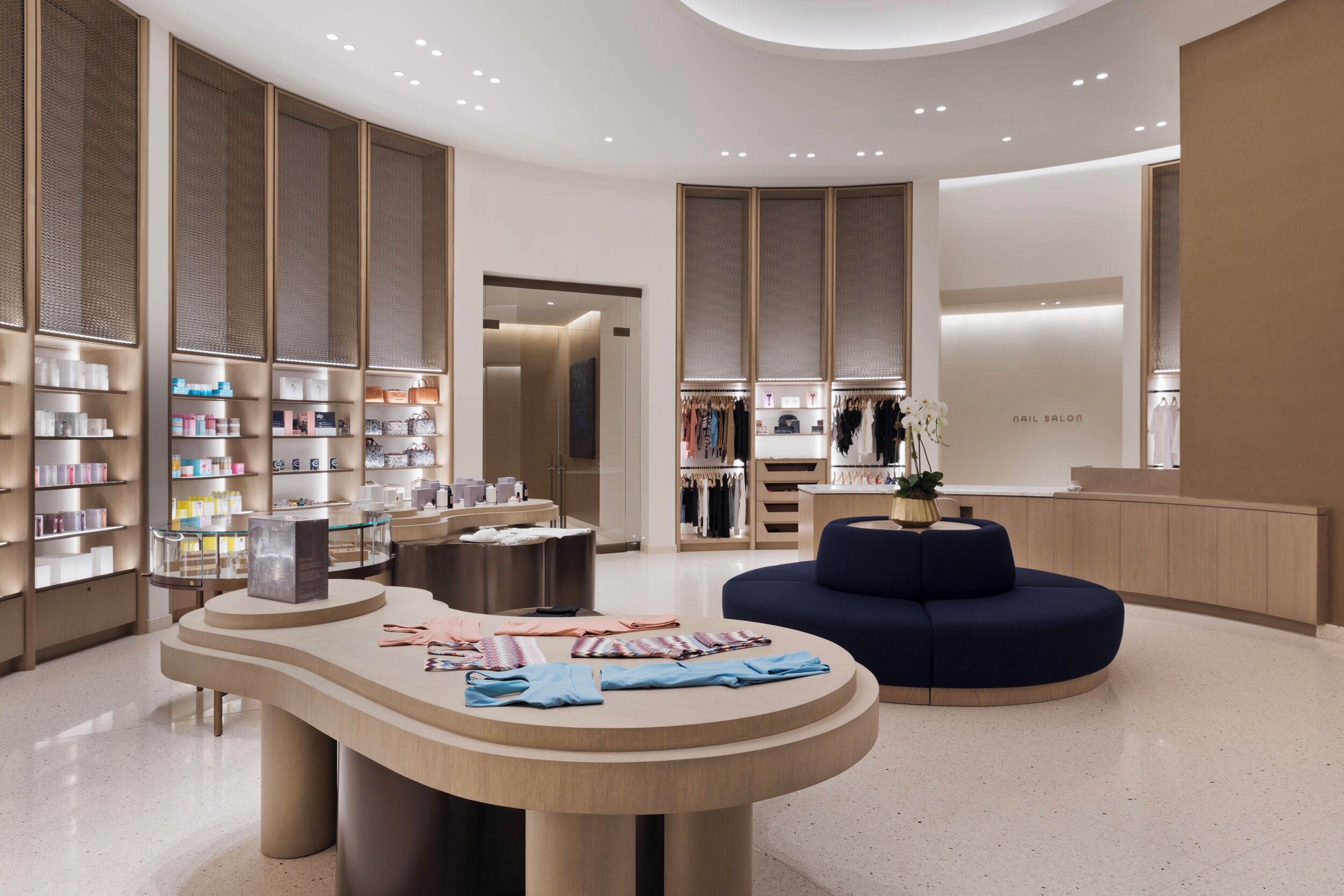 A modern, well-lit retail space with a central round table displaying folded clothes, surrounded by shelves stocked with various products.