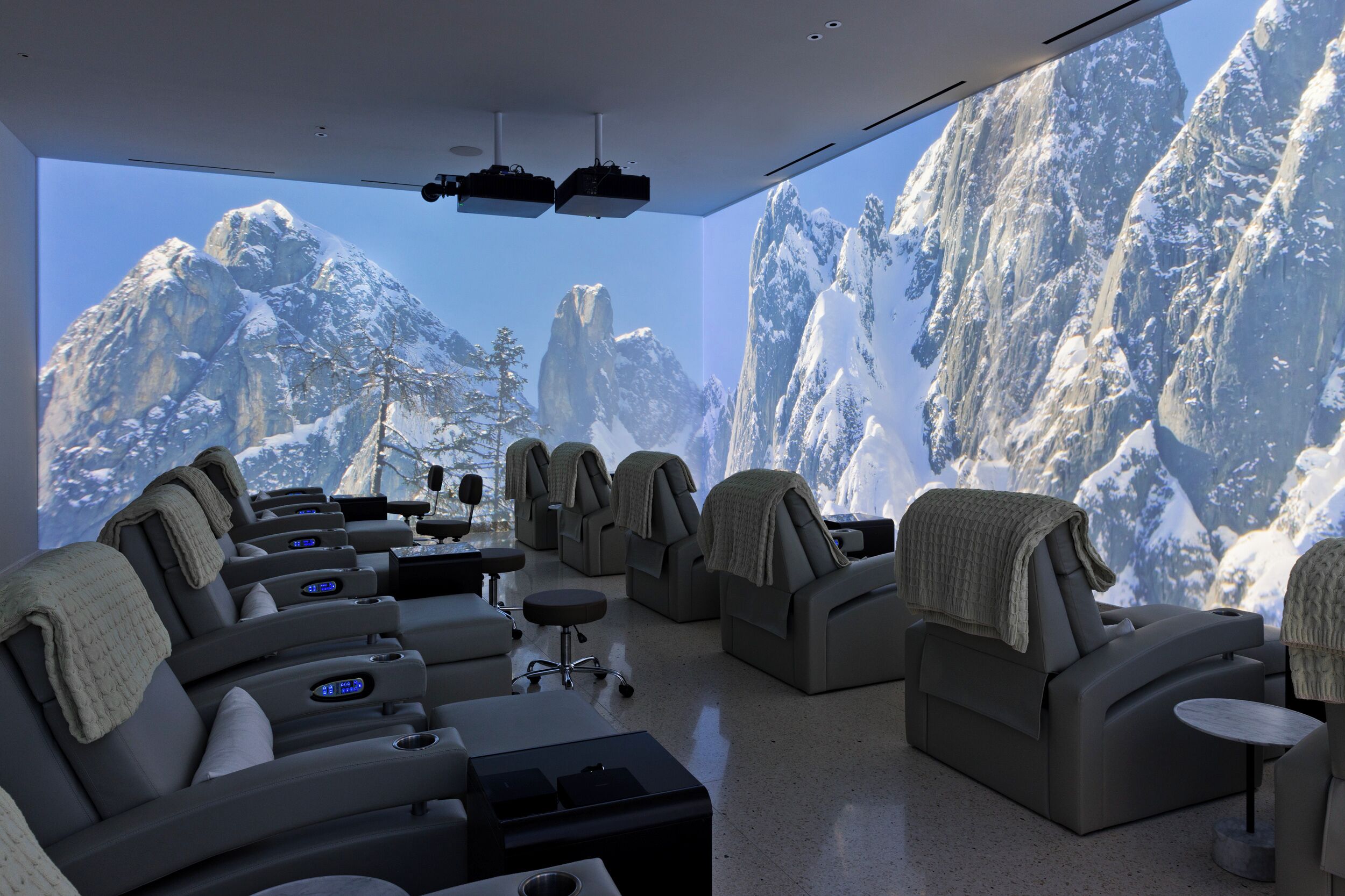 Una sala de relajación moderna con sillones reclinables, una mesa de masajes y una pantalla grande que muestra un paisaje nevado de montañas.