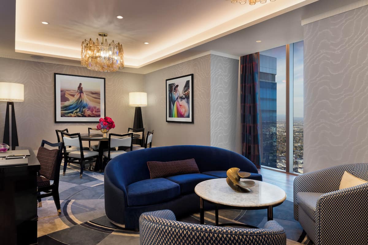 Une chambre d’hôtel moderne avec un canapé bleu, une table basse ronde et un coin repas comprenant des chaises et une table. Deux tableaux encadrés ornent les murs, ainsi qu’un lustre et une grande fenêtre offrant une vue sur la ville.