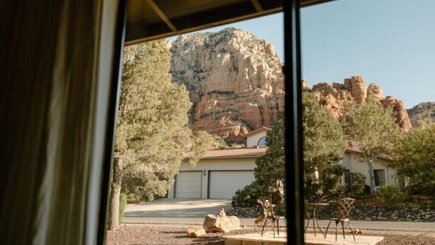 Une vue à travers la fenêtre d’un bâtiment doté d’un garage, d’une terrasse avec des chaises et d’un paysage montagneux.