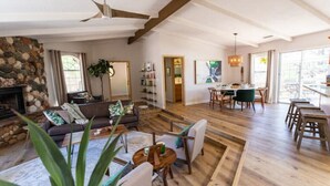 Uma sala de estar ampla com lareira de pedra, piso de madeira, área de jantar com mesa e cadeiras, e uma grande janela que oferece iluminação natural.