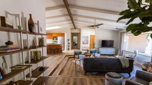 Uma sala de estar ampla com piso de madeira, um sofá escuro e uma televisão. Há prateleiras com livros e objetos decorativos, e uma área de cozinha é visível ao fundo.