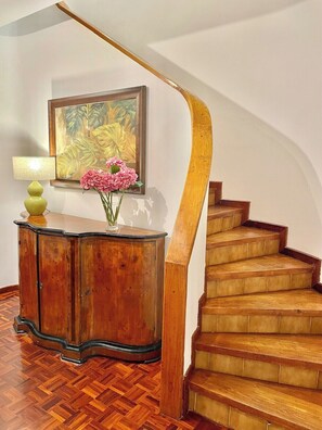 Una escalera de madera con barandilla curva, un mueble de madera con una lámpara y flores, y un cuadro en la pared.