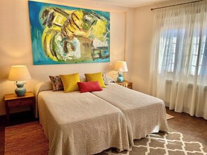 A hotel room with two beds, a large abstract painting on the wall, and a window with sheer curtains.