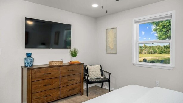 Ein Schlafzimmer mit einem Bett, einem Schminktisch, einem Stuhl, einem Deckenventilator, einem Fernseher und einem Fenster mit Blick auf Grünflächen.