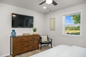 Un dormitorio con cama, cómoda, silla, ventilador de techo, televisor y ventana con vistas a follaje.