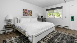 A bedroom with a white bed, a black and white rug, a nightstand with a lamp, a window with a view, and a green door. - Peninsula One Carefree Checkout (Traverse City)