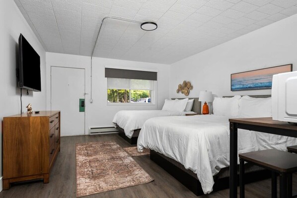 A modern bedroom with two beds, a TV, a wooden dresser, and a window with a view of greenery.
