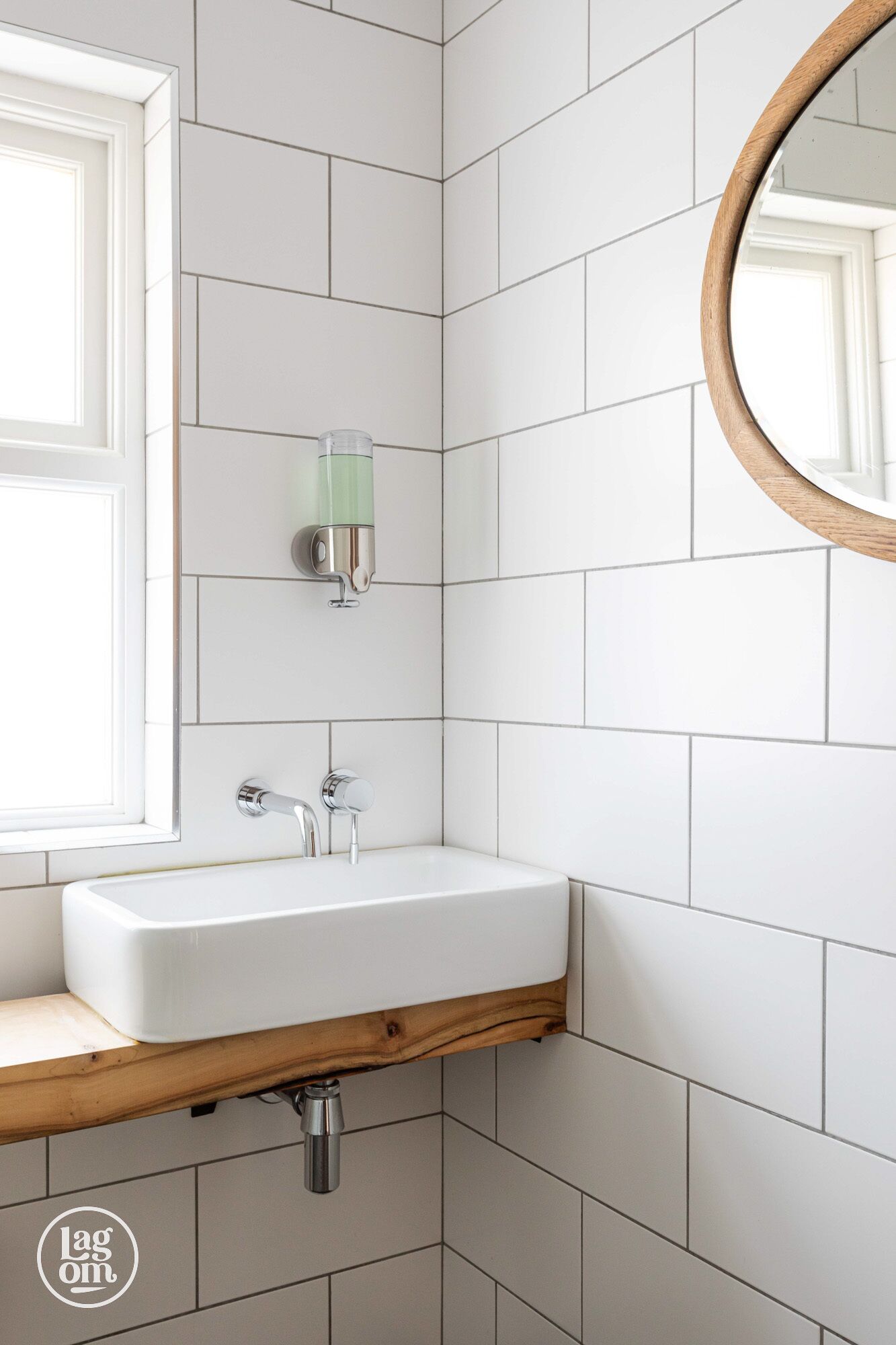 Un baño con pared de azulejos blancos, encimera de madera, lavabo blanco, espejo y dispensador de jabón verde.