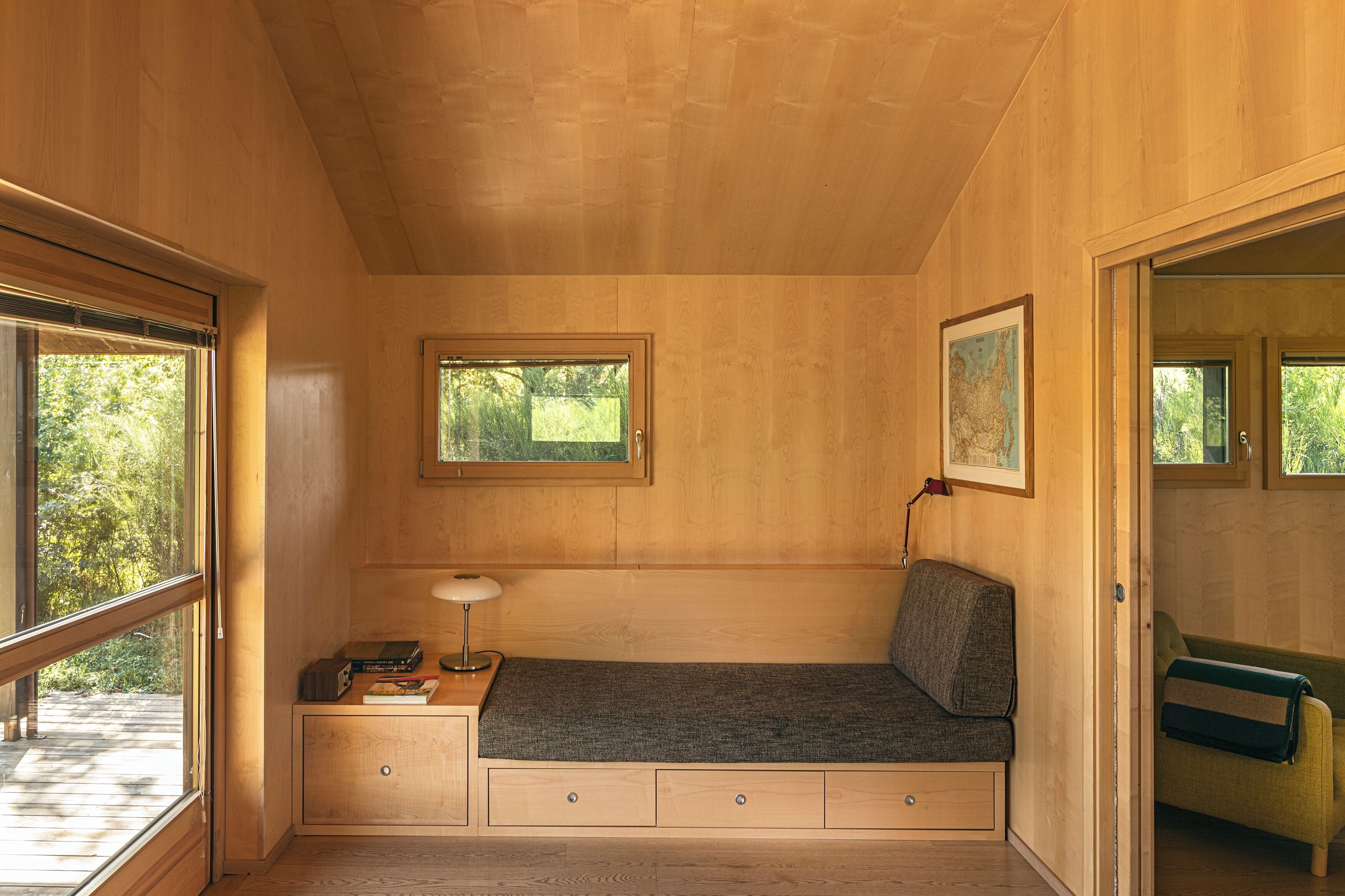 A compact room with a built-in bench, a small desk with a lamp, and a window with a view of greenery.
