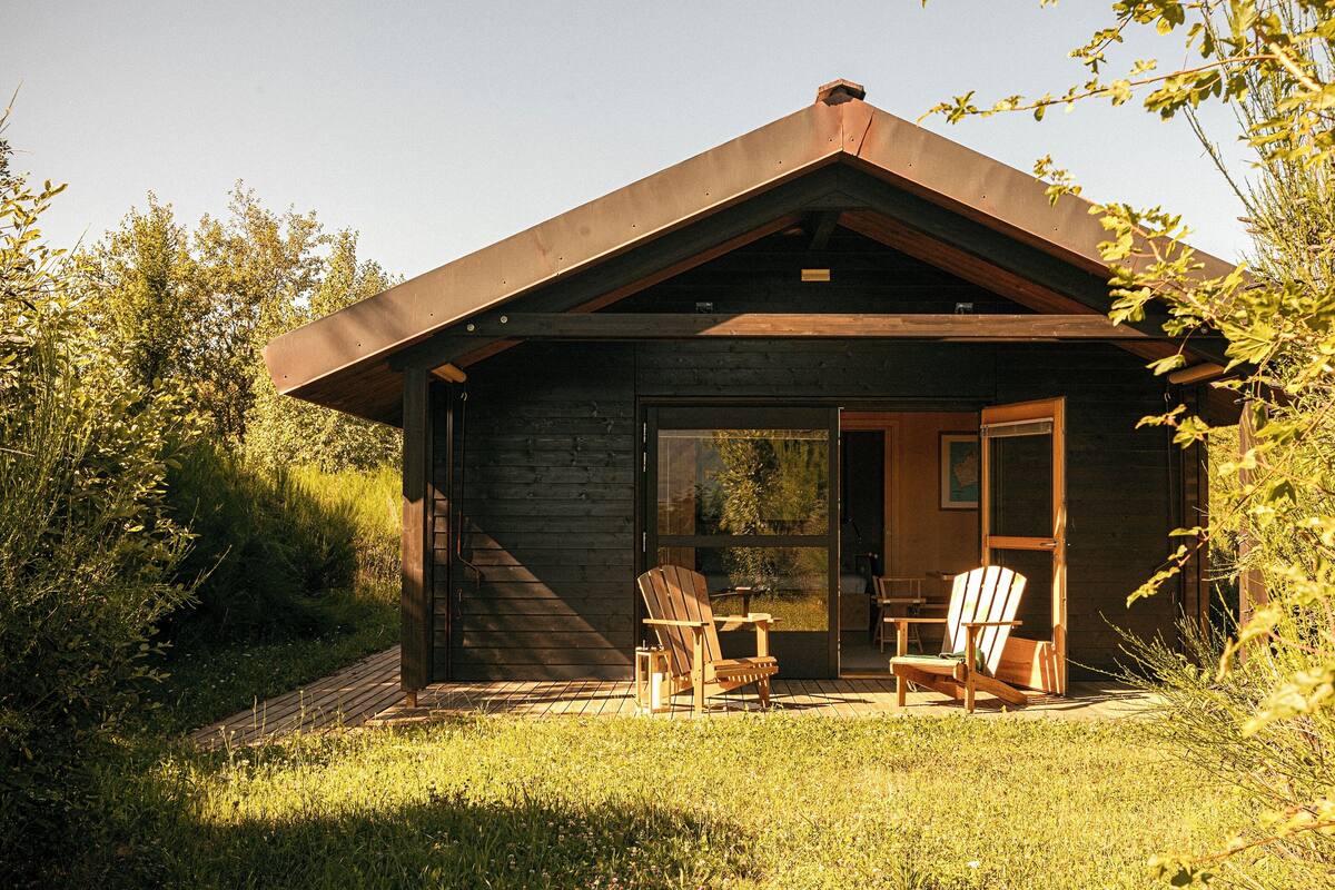 A small wooden cabin with a sloped roof, a glass door entrance, and two wooden chairs outside.