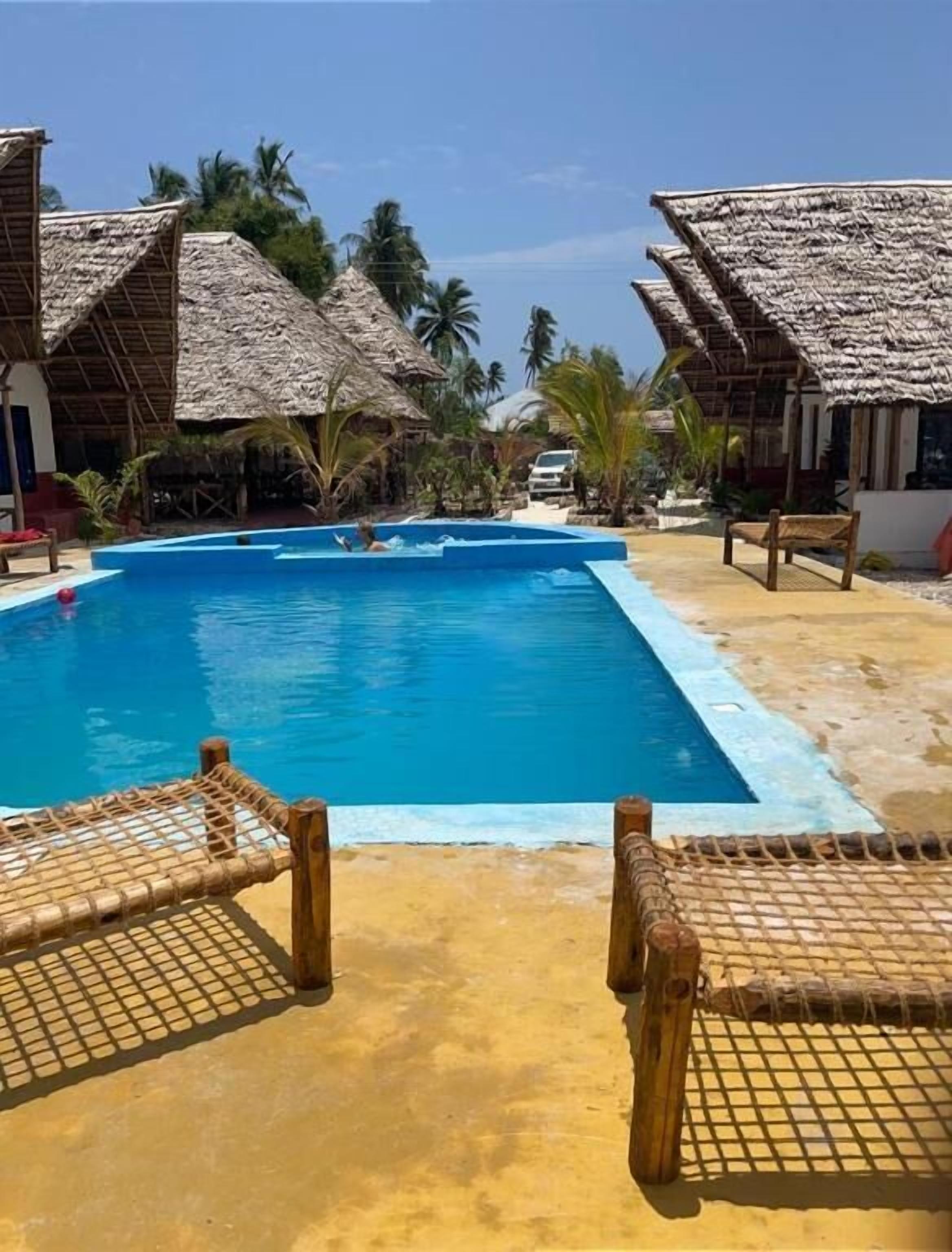 Un área de piscina con estructuras de techo de paja, sillas de descanso y palmeras.