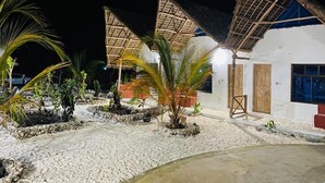 A beachside resort with thatched-roof huts, palm trees, and a sandy pathway leading to a building with a wooden door. - MKEKA SPICE LODGE (Jambiani)