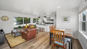 A modern living room with a brown leather sofa, a wooden dining table with chairs, a kitchen area with white cabinets, and large windows offering a view of the outdoors.