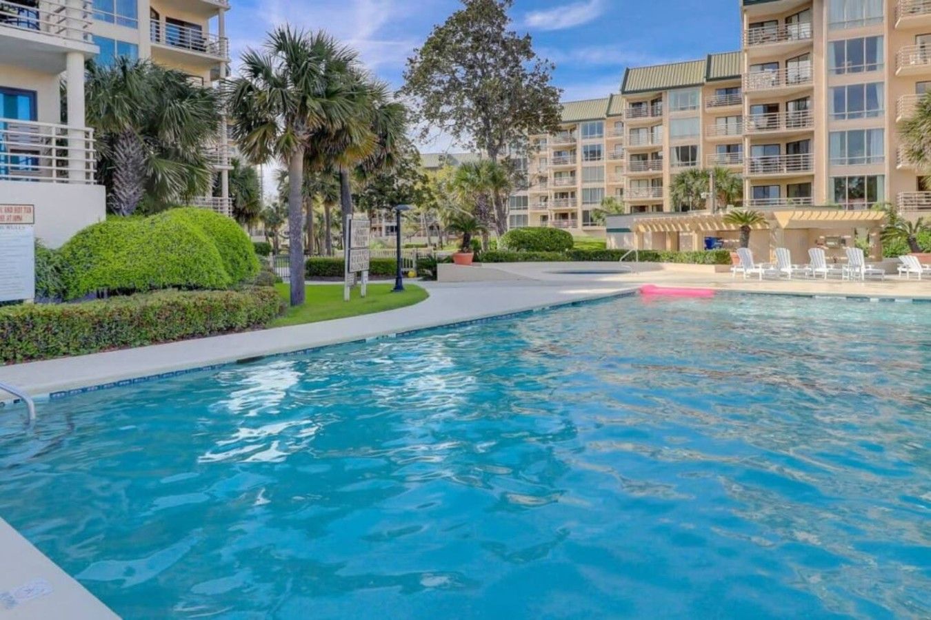 A swimming pool with a clear blue water surface, surrounded by well-maintained greenery and a multi-story building with balconies.