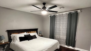 A bedroom with a bed, a nightstand, a lamp, a ceiling fan, and a window with curtains. - 3BR/1BA BROKEN ARROW HOME (Broken Arrow)