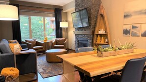 A modern living room with a stone fireplace, a dining area, and a large window offering a view of greenery. - Grotto Mountain Guesthouse (Canmore)