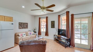Un salon avec un canapé en cuir marron, un téléviseur posé sur un meuble en verre, un canapé beige agrémenté de coussins à motifs floraux, un ventilateur de plafond et un coin cuisine avec un réfrigérateur.