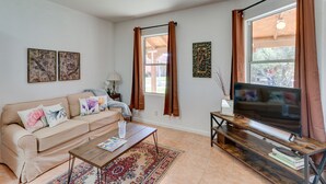Un salon avec un canapé beige, une table basse, un téléviseur posé sur un meuble en bois et une grande fenêtre avec des rideaux.