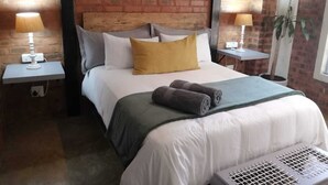 A hotel room with a brick wall, a bed with white bedding and a mustard-colored pillow, two bedside tables with lamps, and a potted plant.