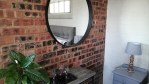 A room with a brick wall, a round mirror, a bedside table with a lamp, and a potted plant.