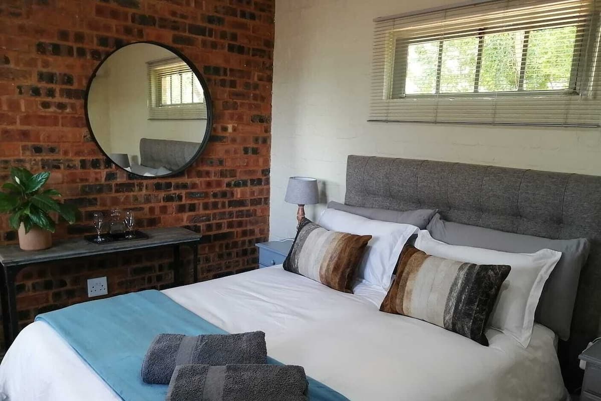 A bedroom with a brick wall, a round mirror, a bed with multiple pillows, a bedside table with a lamp, and a window with blinds.