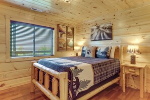A wooden cabin bedroom with a bed, a nightstand, a lamp, and a window with blinds. - Gorgeous Gatlinburg Cabin: Large Deck & Hot Tub! (Gatlinburg)