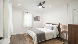 Ein Schlafzimmer mit Bett, Deckenventilator, Nachttisch mit Lampe und einem Fenster mit Vorhängen.