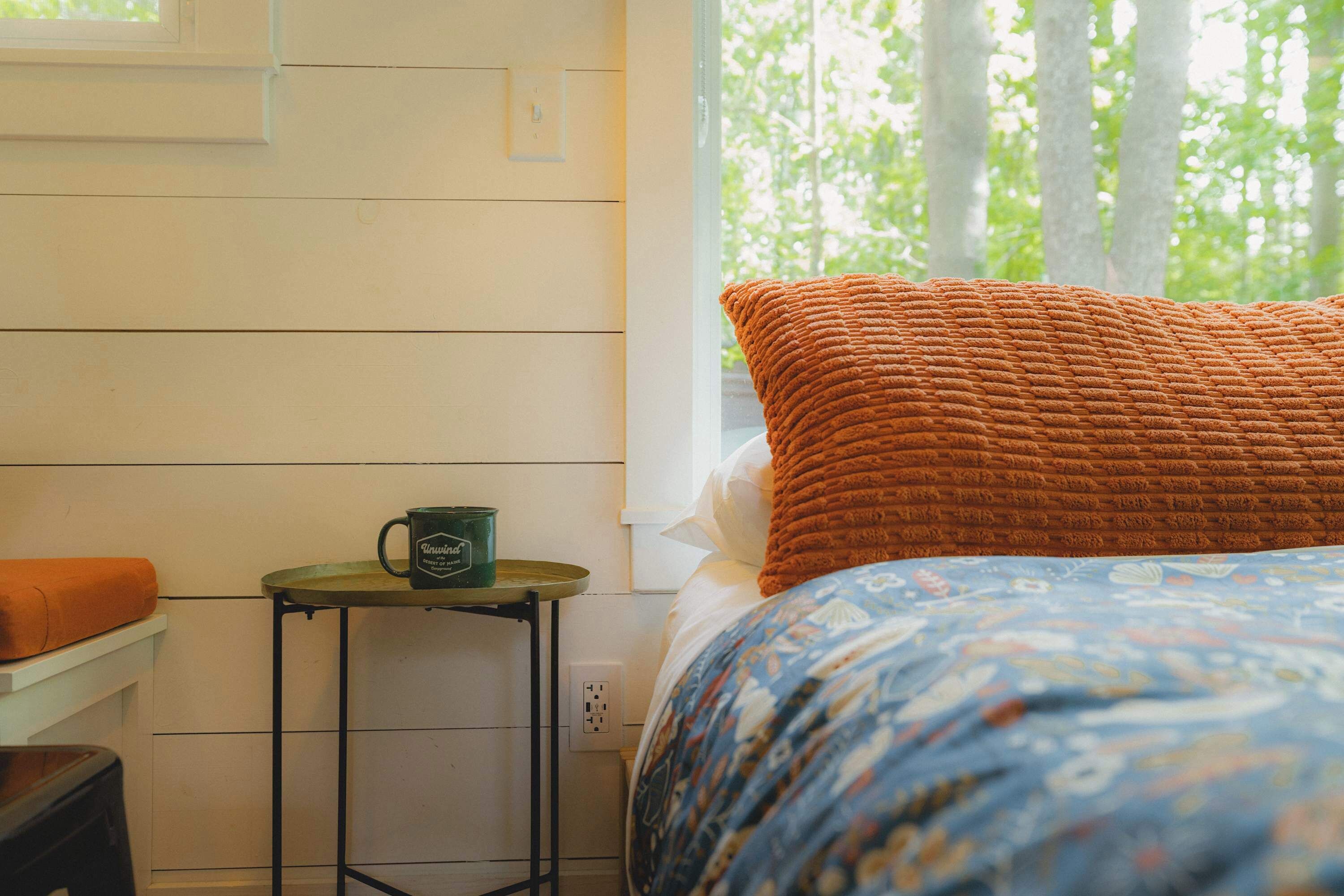 A bedroom with a bed, a window with a view of trees, a small table with a mug, and a chair.