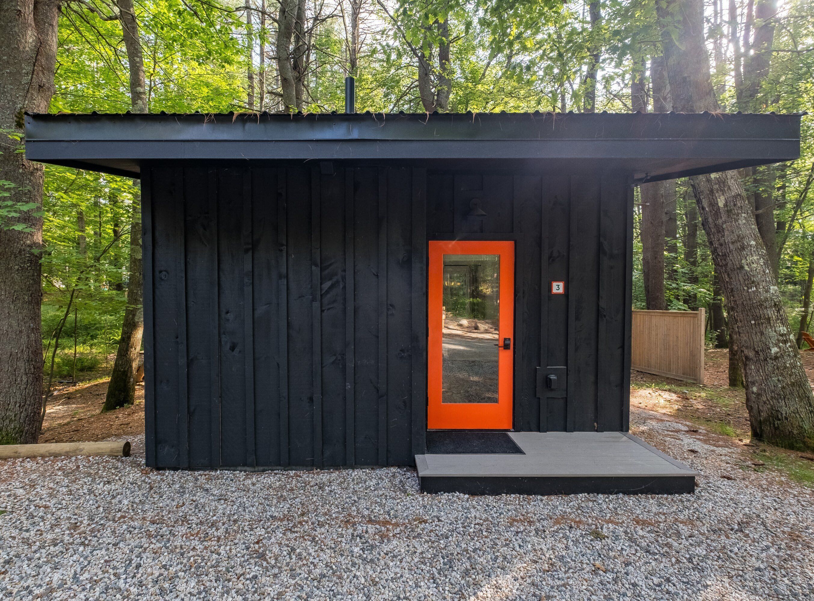 A small, dark wooden cabin with a bright orange door, surrounded by trees.