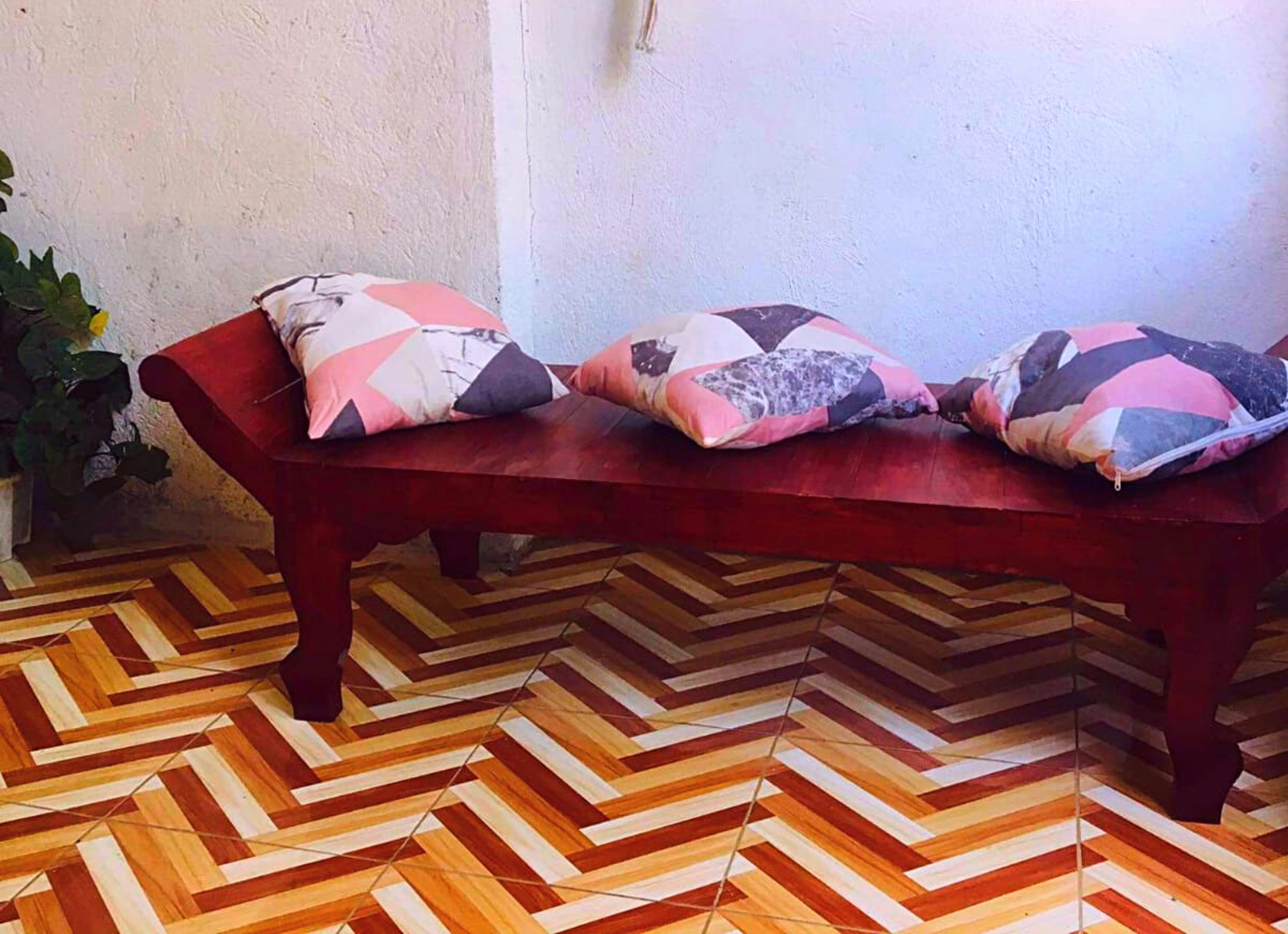 A wooden bench with patterned cushions against a textured wall, a hanging plant, and a herringbone-patterned floor.