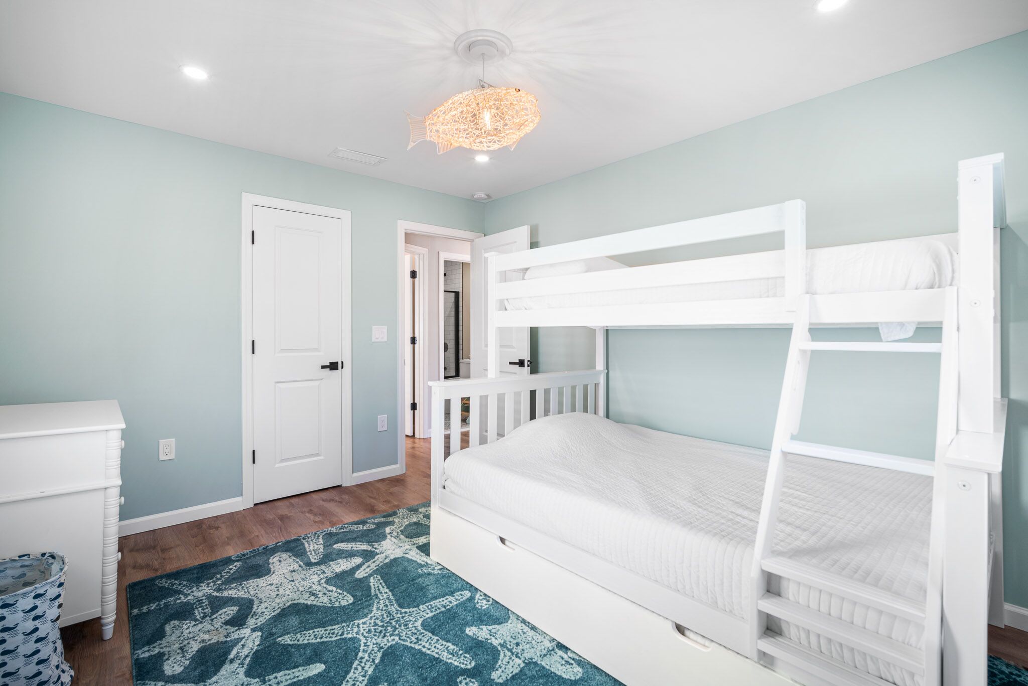 Une chambre avec des lits superposés, une commode blanche, un tapis orné de motifs d’étoiles de mer et un luminaire au plafond.