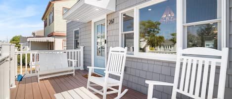 A wooden deck with white rocking chairs and a bench, a large window with a view of a building, and a number '261' on the wall.