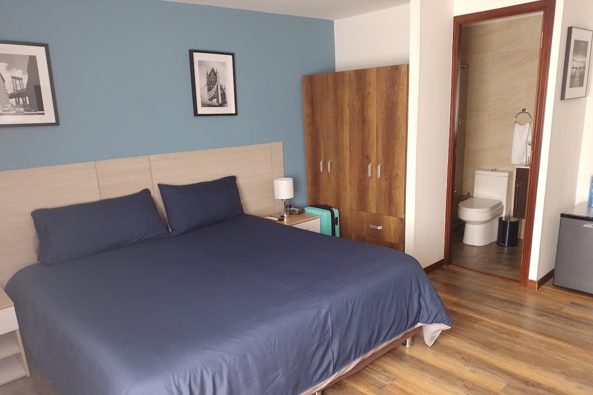 Een netjes opgemaakt bed met een blauwe spreidekker, een houten kast, een klein nachtkastje met een lamp en een lijst met een foto aan de muur.