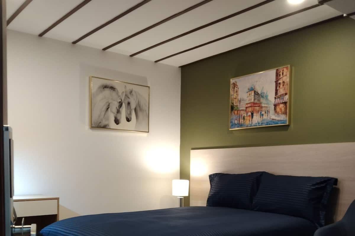 Een slaapkamer met een groot bed, een hoofdbord, een bureau, een stoel, een lamp en twee schilderijen aan de muur.
