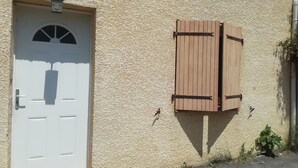 A building with a textured beige exterior, a white door with a small window, and a wooden shutter. - Maison à la Campagne (Corneilhan)