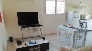A living room with a TV on a stand, a sofa, a coffee table with a remote and tissues, and a kitchen area with a fridge, microwave, and dining table. - Maison à la Campagne (Corneilhan)