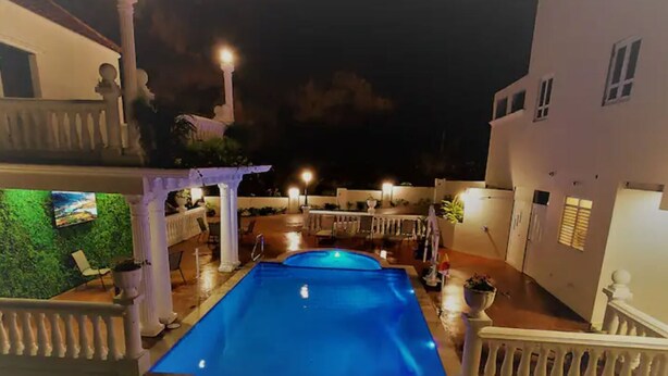 Una piscina con una pérgola y zona de estar al aire libre, rodeada por una cerca blanca y un edificio con balcón.