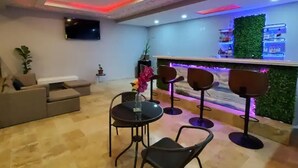 Un área de estar moderna con un sofá, una mesa redonda con sillas, un televisor montado en la pared y una zona de bar con pared verde e iluminación azul.