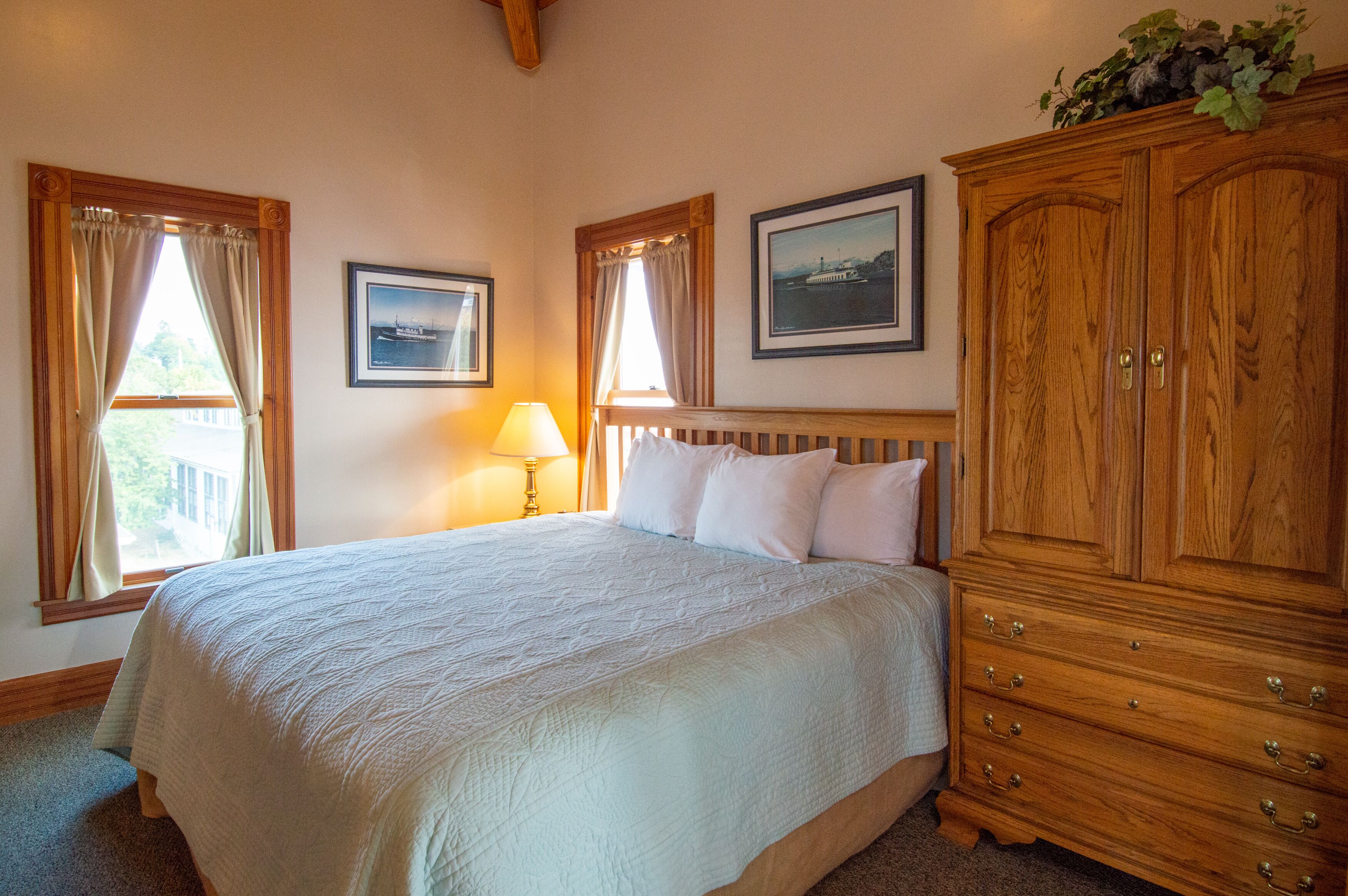 Une chambre avec un grand lit, une armoire en bois, deux fenêtres avec des rideaux, une lampe et des tableaux encadrés accrochés au mur.