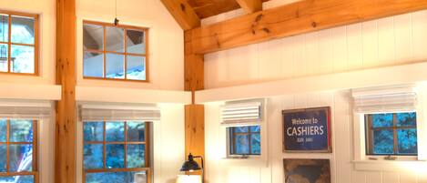 A spacious room with a high ceiling, wooden beams, a ceiling fan, a brown leather sofa, a wooden coffee table, and a sign reading "Welcome to CASHIERS ELEV. 3481".