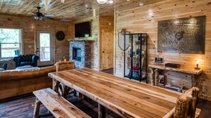 L'intérieur d'un chalet en bois comprend une grande table à manger, une cheminée, un canapé en cuir et une carte accrochée au mur.