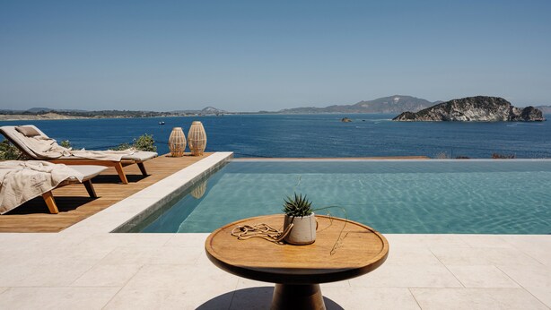 Zona junto a la piscina con sillones de descanso y una mesa, con vistas al mar azul y montañas a lo lejos.