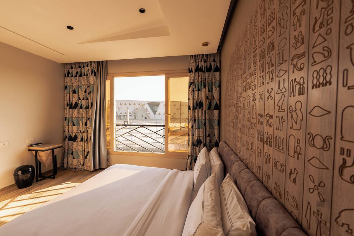 A hotel room with a large bed, a small table, and a view of a building through the window.
