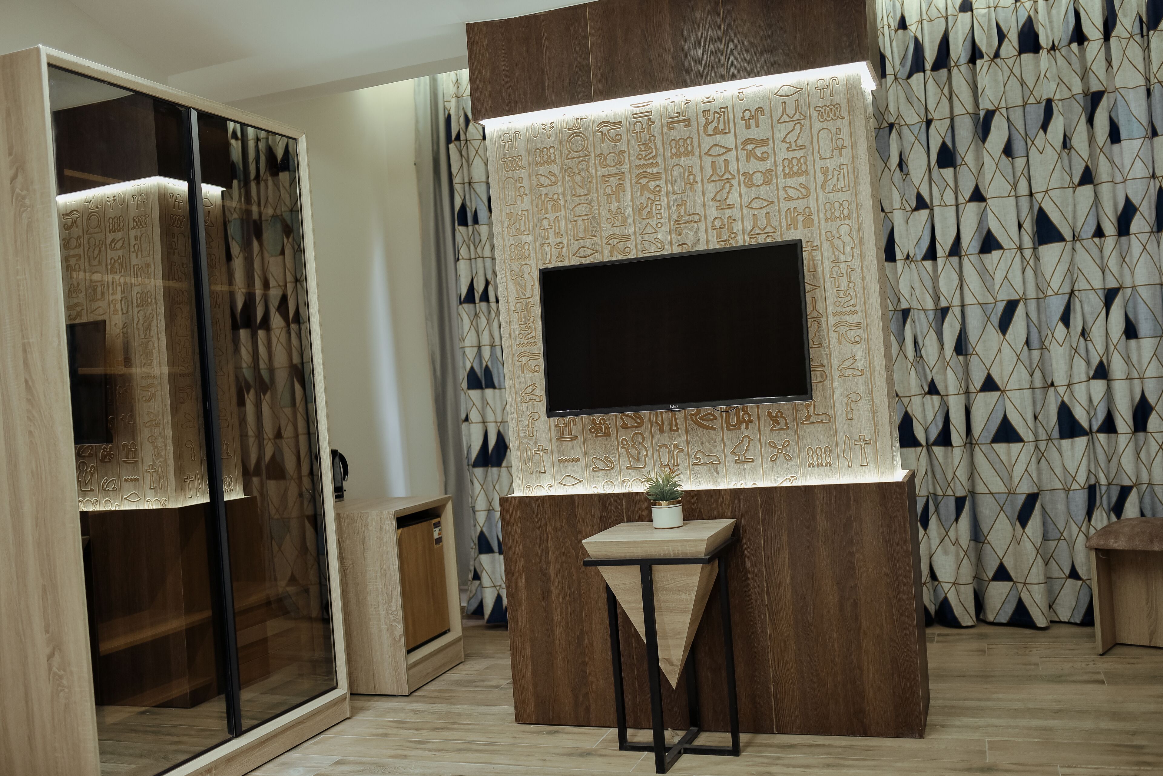 A modern living room with a large flat-screen TV mounted on a wooden wall, a small wooden table with a plant, and a patterned curtain.