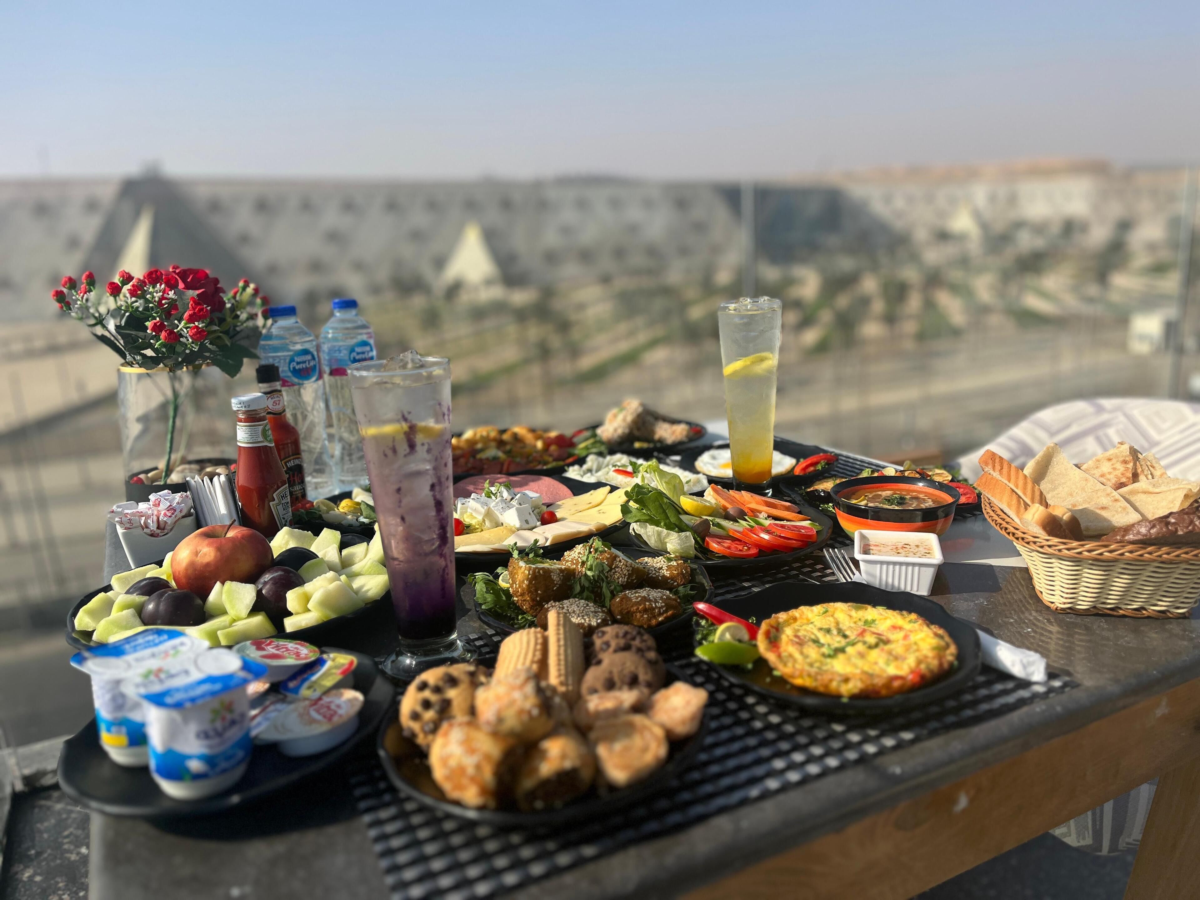 Ein Tisch mit einer Auswahl an Speisen und Getränken, darunter Früchte, Käse und ein Salat, vor einer schönen Aussicht.