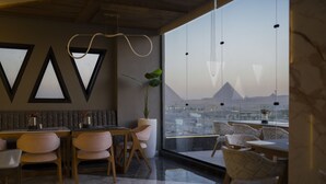 A modern dining area with a city view, featuring a long table with chairs, a potted plant, and geometric wall decorations. - Royal Great Pyramid INN (Giza)